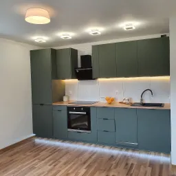 Nowoczesna kuchnia z zielonymi szafkami, drewnianym blatem, białym marmurowym backsplashiem, oświetleniem LED pod szafkami i drewnianą podłogą.