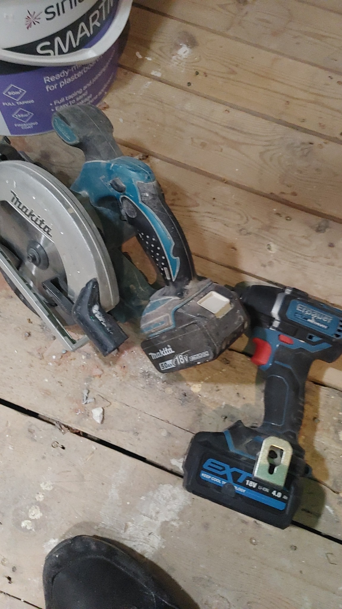 Używane narzędzia budowlane: piła tarczowa Makita, wiertarka Makita i wkrętarka Erbauer, leżące na drewnianej podłodze obok wiadra z gotową masą szpachlową.