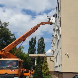 Montaż lub naprawa osłon okiennych na elewacji budynku z użyciem podnośnika koszowego; pracownik w koszu na wysokości okna.