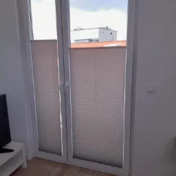 Zamknięte plisowane osłony okienne w białej ramie okiennej, widok z wnętrza pomieszczenia na budynek na zewnątrz.