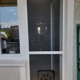Biała rama drzwi balkonowych z siatką i klapką dla zwierząt z odciskiem łapy w dolnej części, widok na zewnątrz przez okno obok.