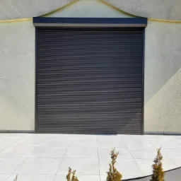 Ciemnoszara roleta zewnętrzna opuszczona na parterze domu z jasną elewacją, widok z poziomu wypielęgnowanych iglaków.