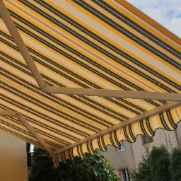 Rozłożona markiza w żółto-czarne pasy, zamontowana nad oknem domu, widok od dołu z fragmentem ogrodu w tle.