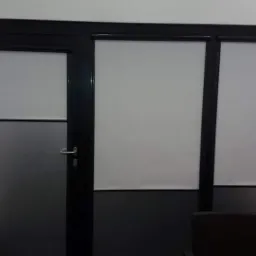 Zestawienie drzwi z matową szybą i trzema oknami z roletami wewnętrznymi w czarnych ramach, widoczne dwa krzesła.