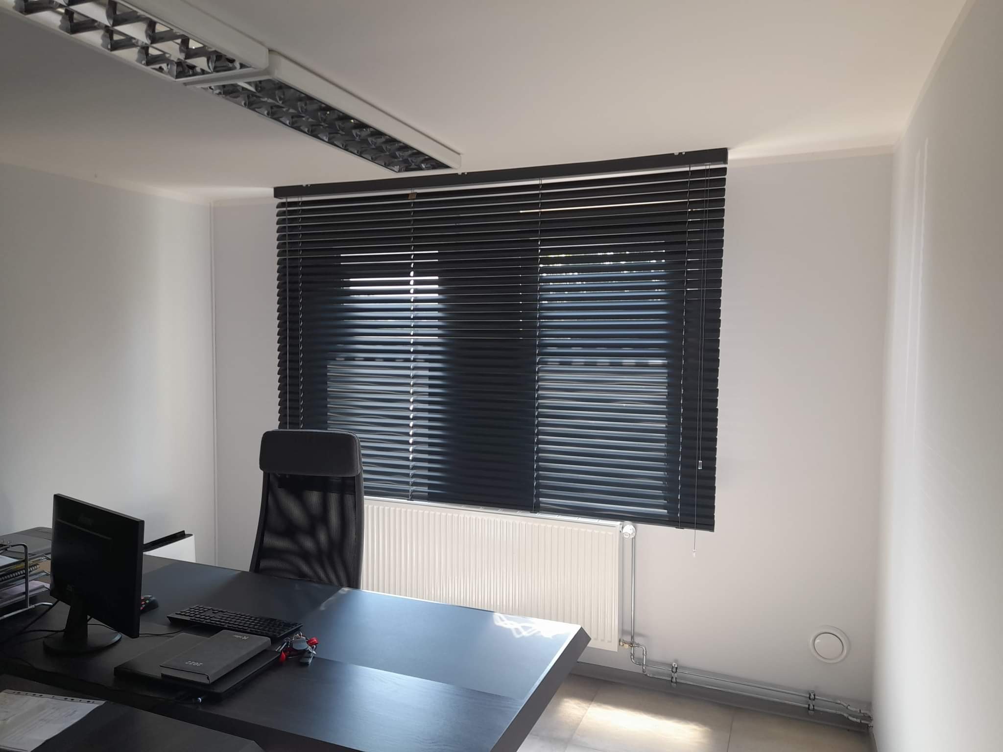 Biuro z czarnymi żaluzjami aluminiowymi na oknie, biały grzejnik pod oknem, czarne biurko z komputerem i krzesłem.