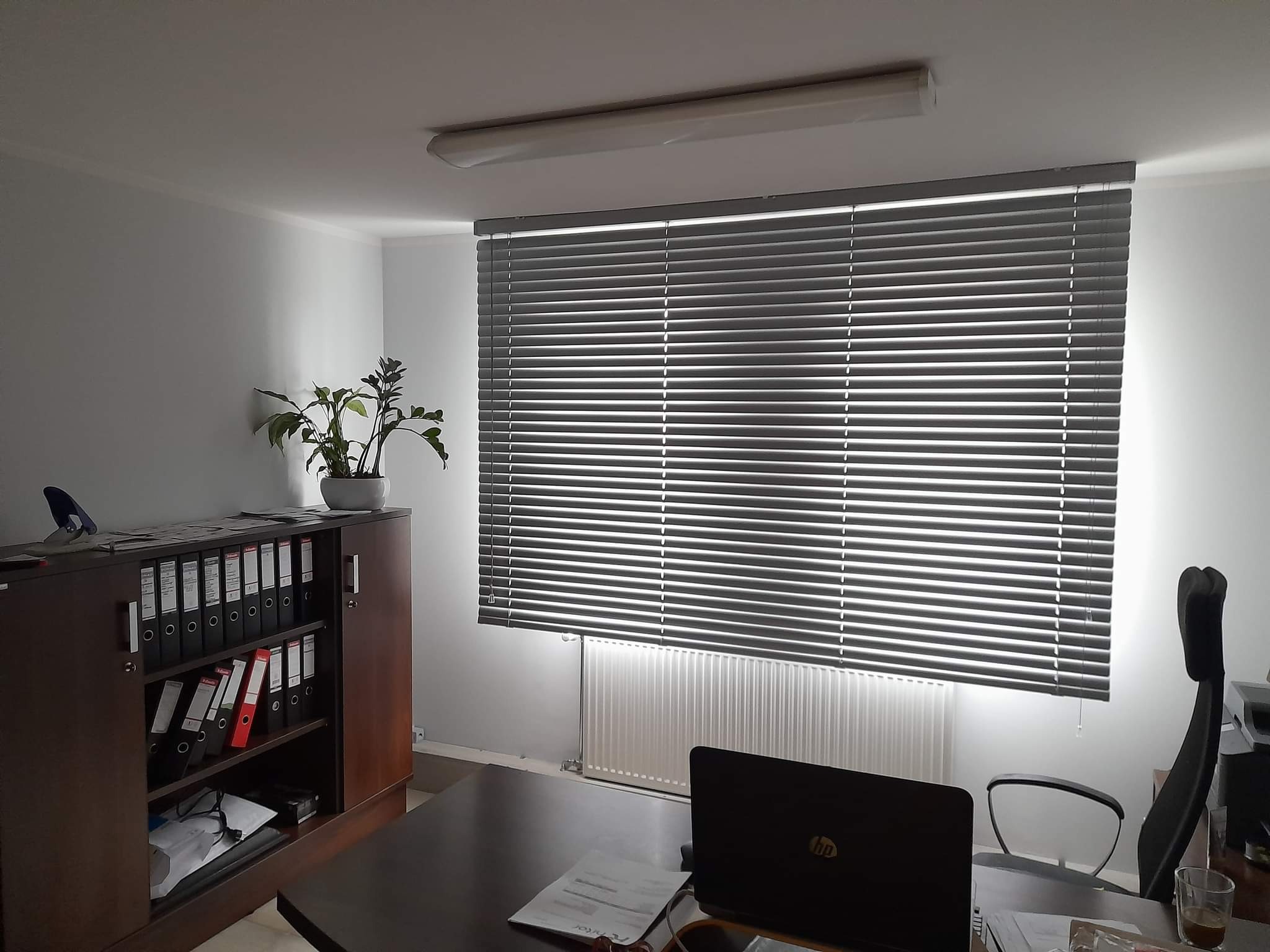 Szare poziome listwy osłaniają okno w biurze, za biurkiem widoczny laptop, obok regał z segregatorami i doniczką z rośliną.