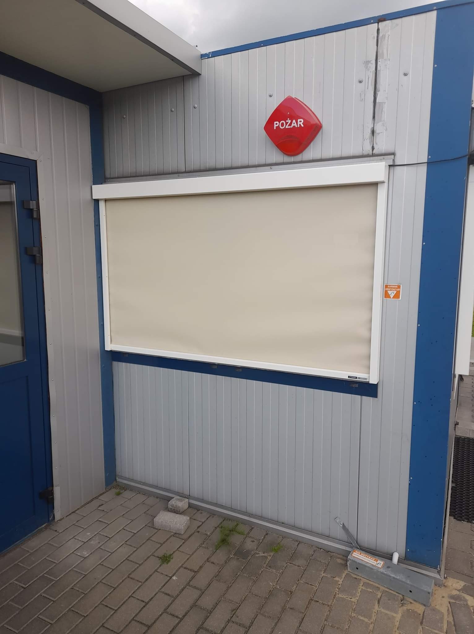 Zamknięta, beżowa przesłona okienna w białej ramie na szarej ścianie kontenera z niebieskimi akcentami, widoczny czujnik pożarowy i kostka brukowa.