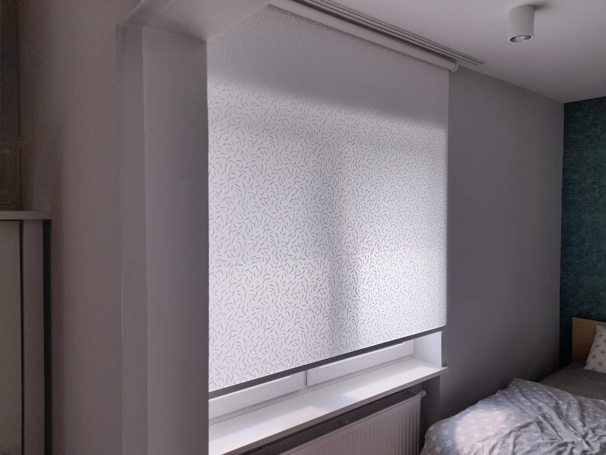 Zamknięta roleta wewnętrzna z delikatnym, roślinnym wzorem na oknie w pokoju, widoczny grzejnik pod parapetem.