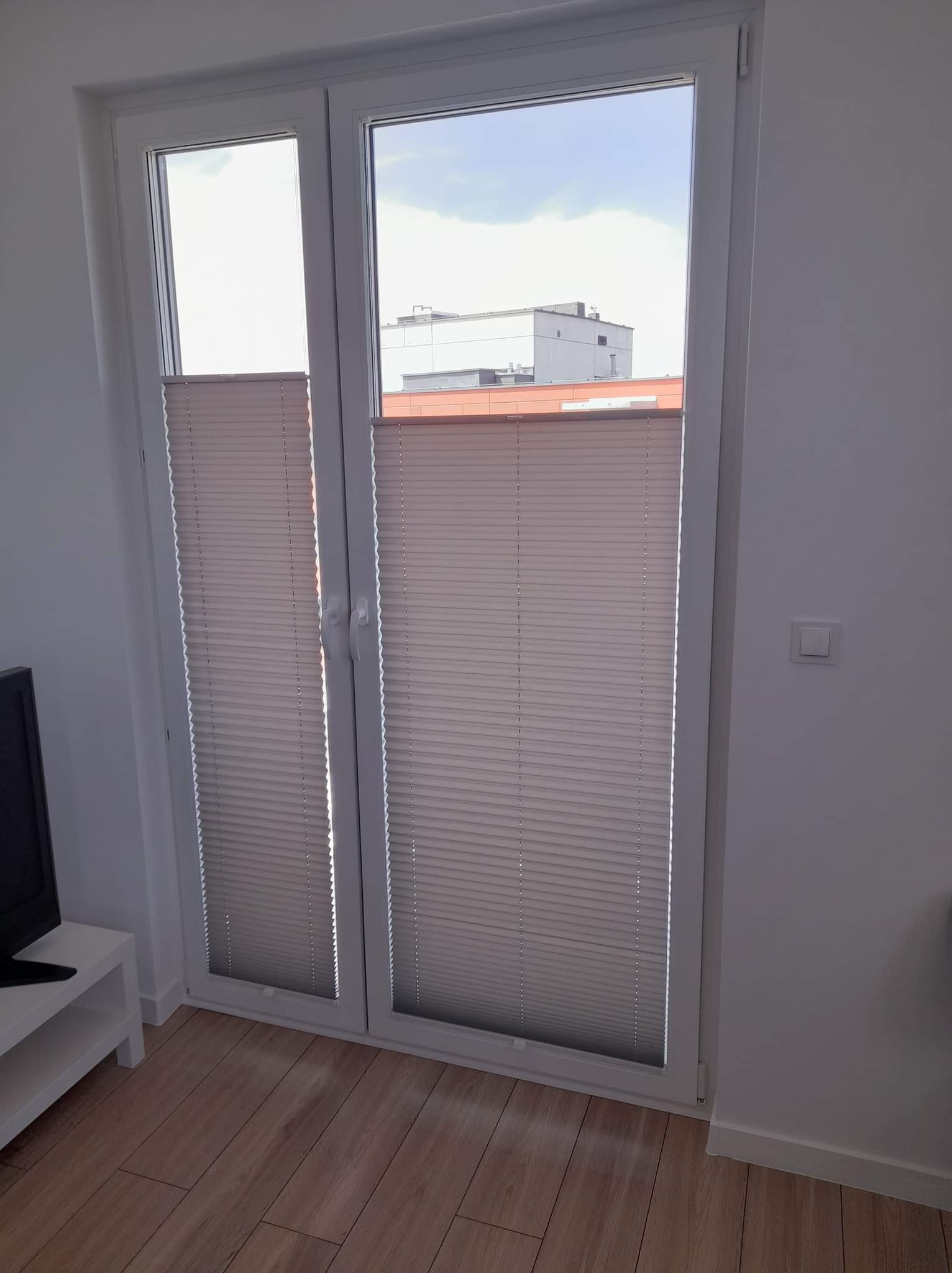 Zamknięte plisowane osłony okienne w białej ramie okiennej, widok z wnętrza pomieszczenia na budynek na zewnątrz.