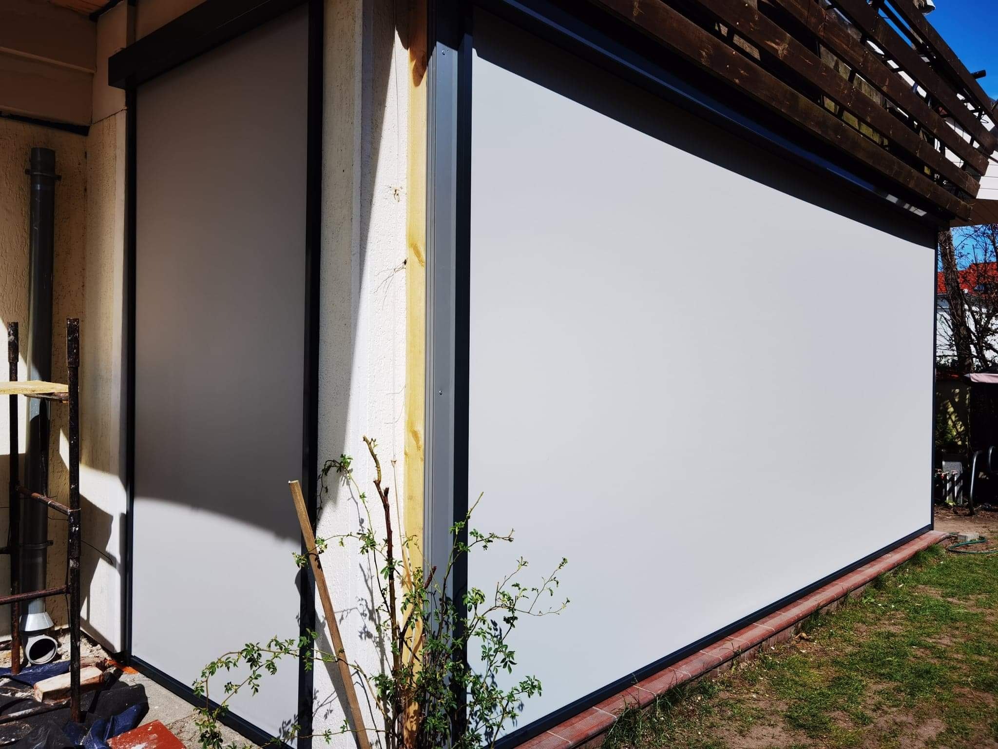 Zewnętrzna przesłona okienna w kolorze białym z czarną ramą, zamontowana na elewacji domu, widok z boku, z zielenią w dolnej części kadru.