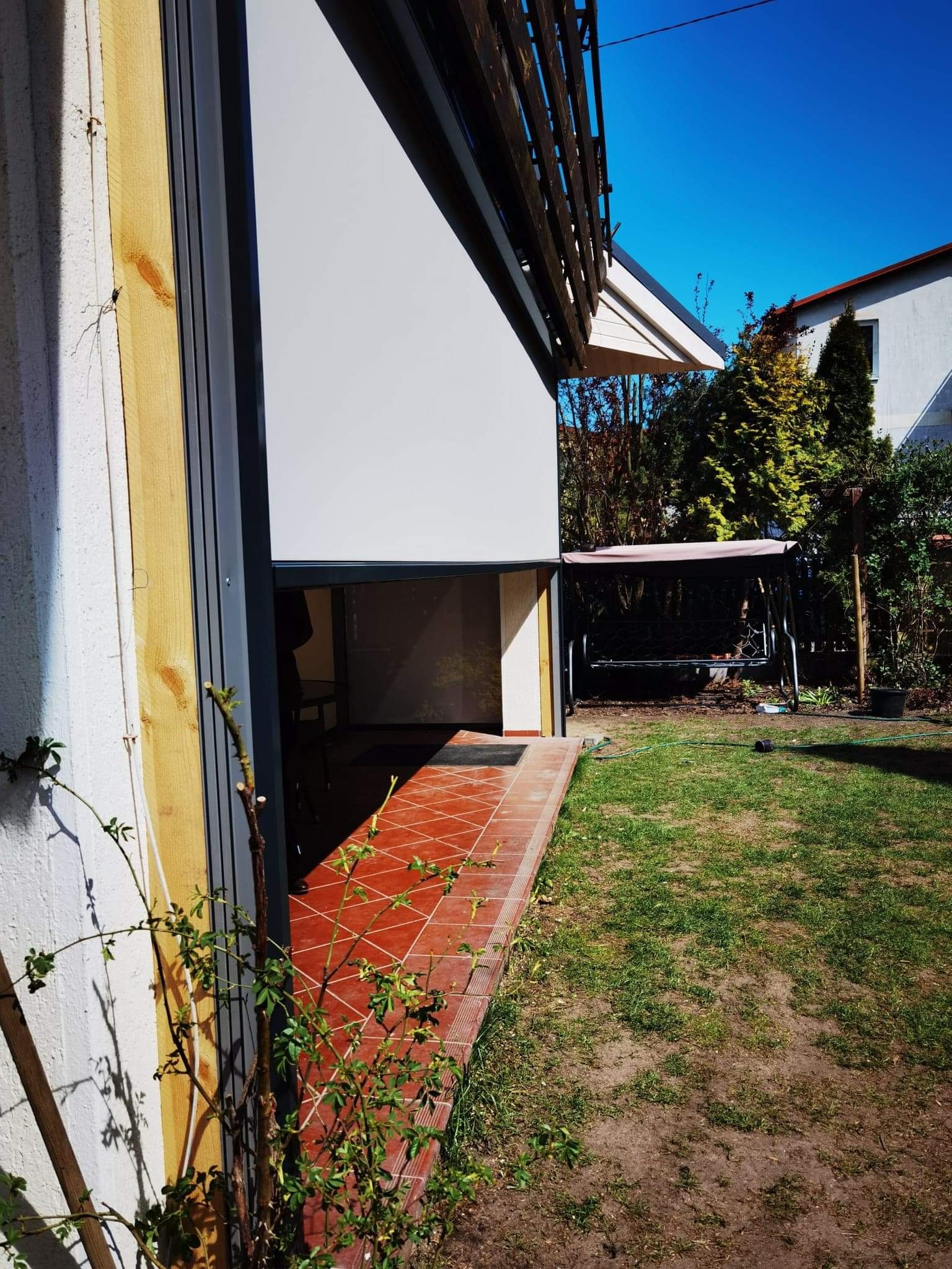 Biała roleta zewnętrzna częściowo zasłonięta, zamontowana na tarasie z czerwoną terakotą, widok z ogrodu w słoneczny dzień.