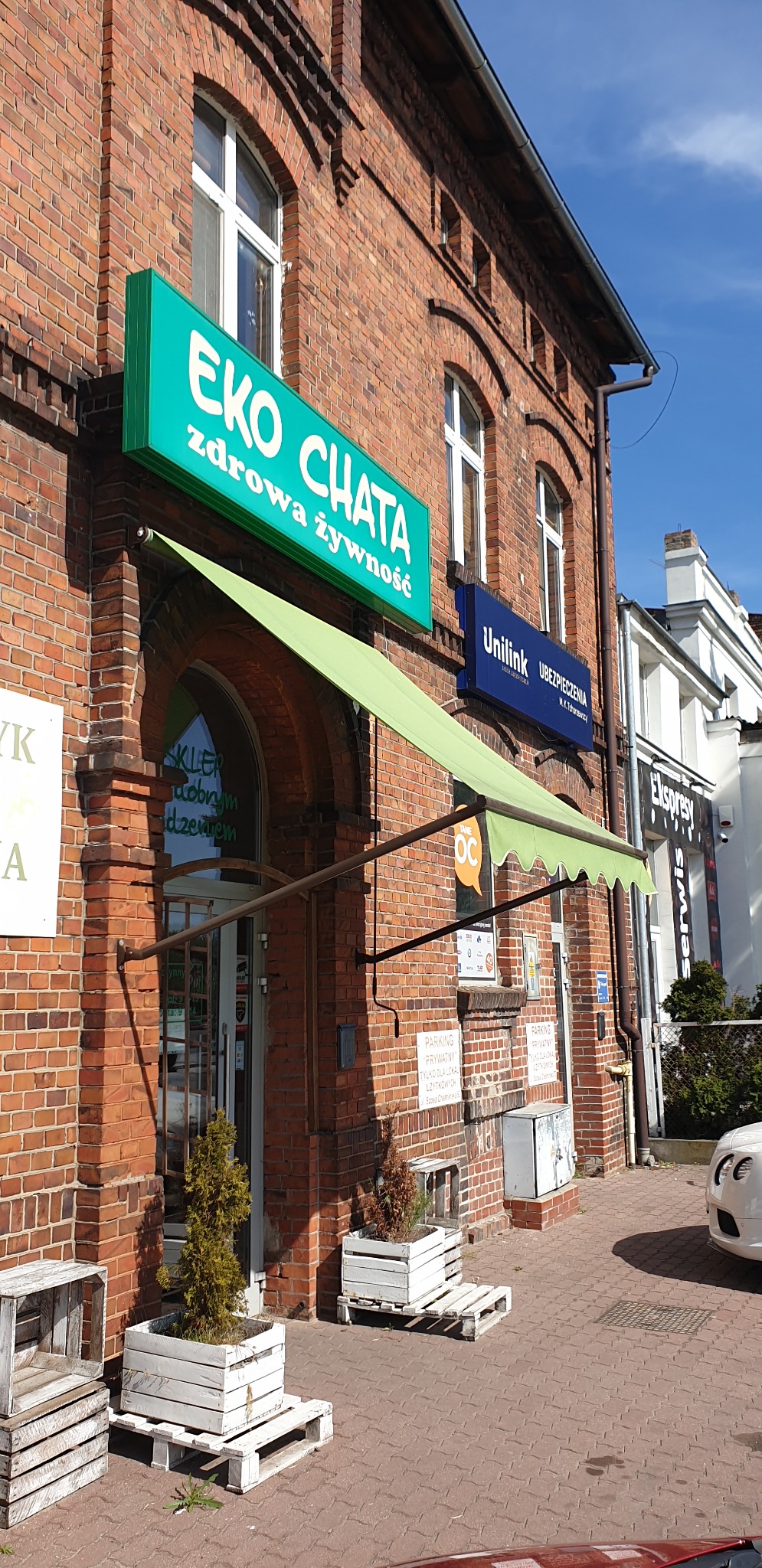 Zielony, rozkładany daszek nad wejściem do sklepu z szyldem 'EKO CHATA zdrowa żywność', fasada z czerwonej cegły, rośliny w donicach na paletach przed wejściem.