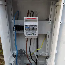 Wnętrze białej skrzynki elektrycznej z zamontowanym zabezpieczeniem przepięciowym marki Apator, widoczne przewody zasilające w różnych kolorach. Zabezpieczenie zamontowane na metalowej szynie DIN.