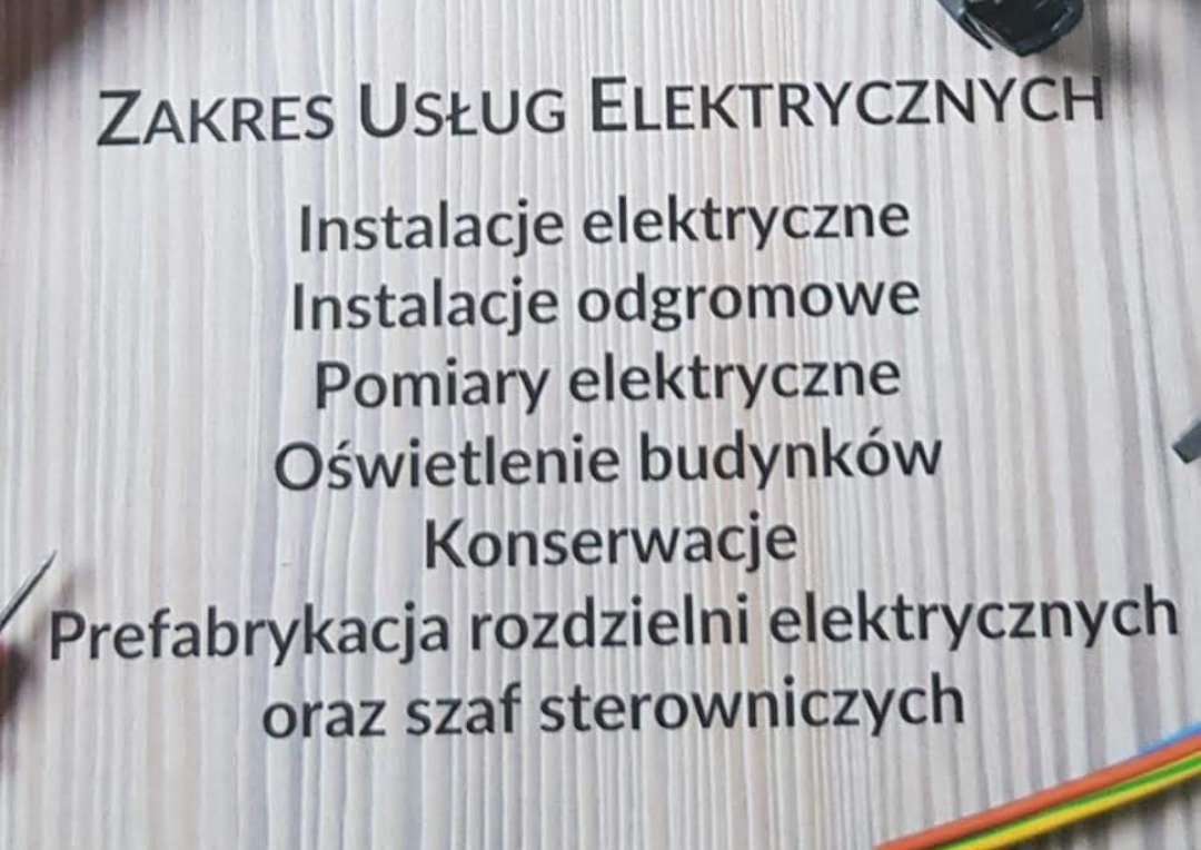 Tablica informacyjna z zakresem usług elektrycznych: instalacje, odgromowe, pomiary, oświetlenie budynków, konserwacje, prefabrykacja rozdzielni i szaf sterowniczych, widoczne fragmenty narzędzi...