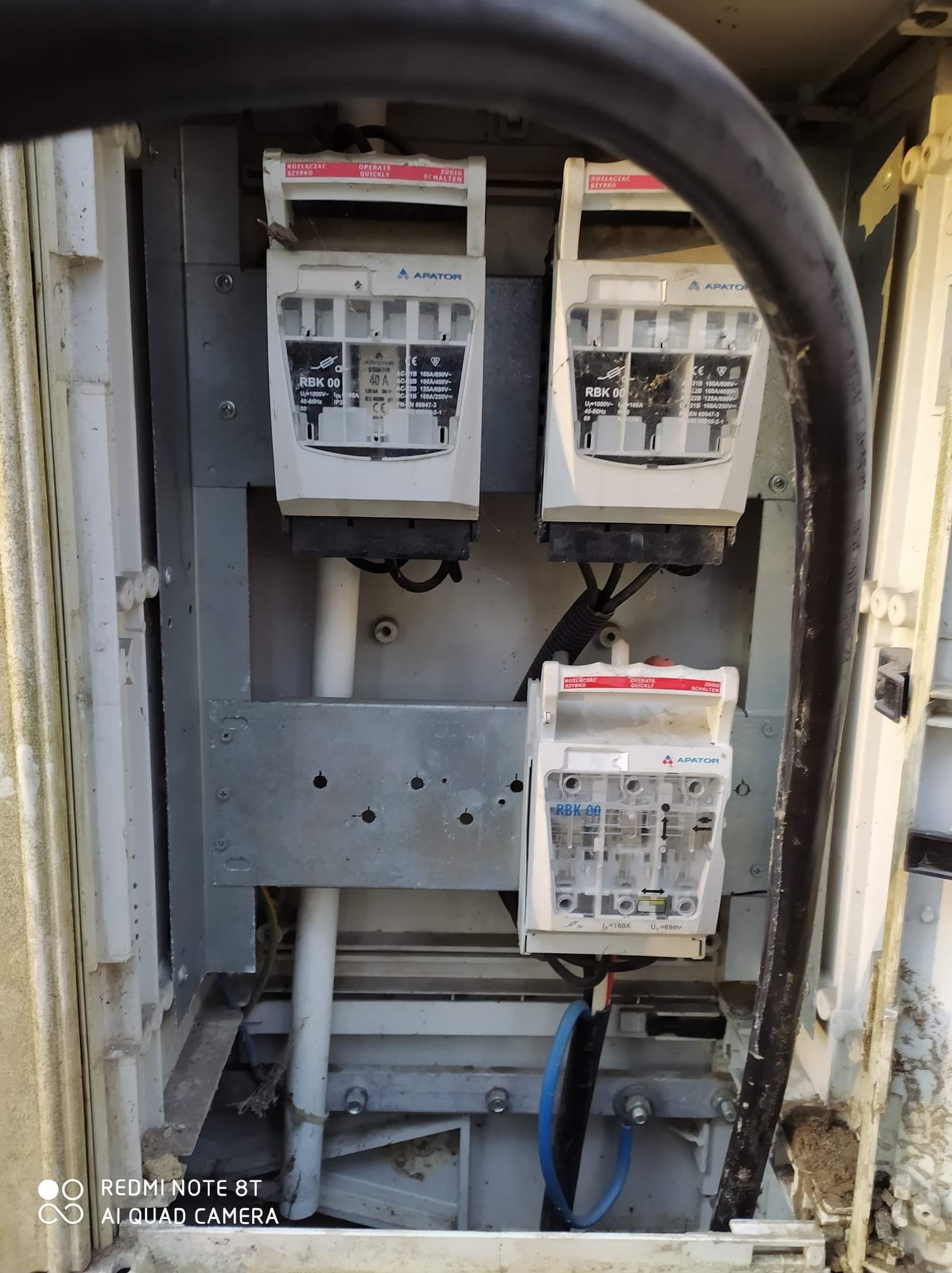 Otwarta skrzynka elektryczna z trzema rozłącznikami bezpiecznikowymi RBK 00 firmy Apator, widoczne kable i przewody, skrzynka brudna i zakurzona.