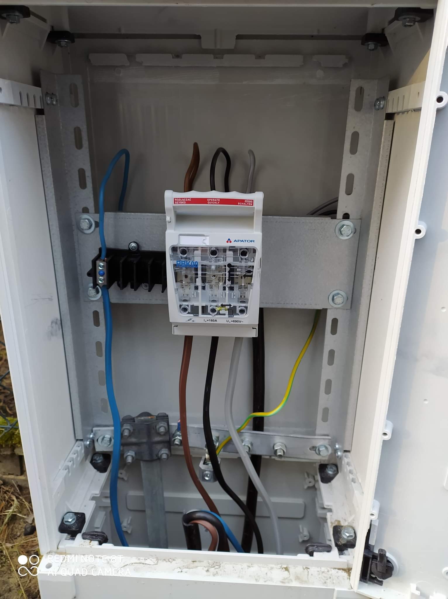 Wnętrze białej skrzynki elektrycznej z zamontowanym zabezpieczeniem przepięciowym marki Apator, widoczne przewody zasilające w różnych kolorach. Zabezpieczenie zamontowane na metalowej szynie DIN.