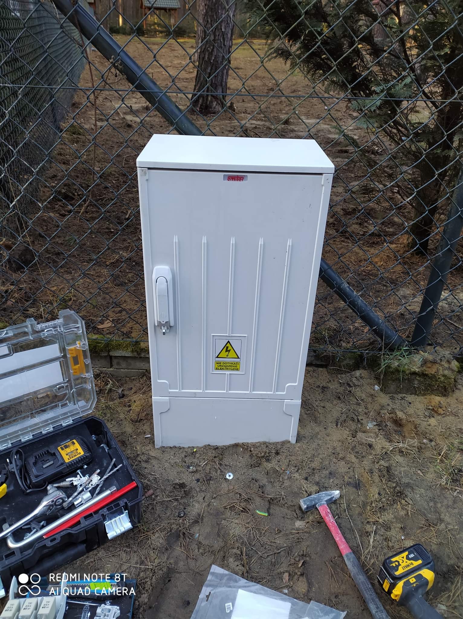 Biała skrzynka elektryczna z symbolem ostrzegawczym na zewnątrz, ustawiona na ziemi przy siatce ogrodzeniowej, obok skrzynki z narzędziami DeWalt i młotka.