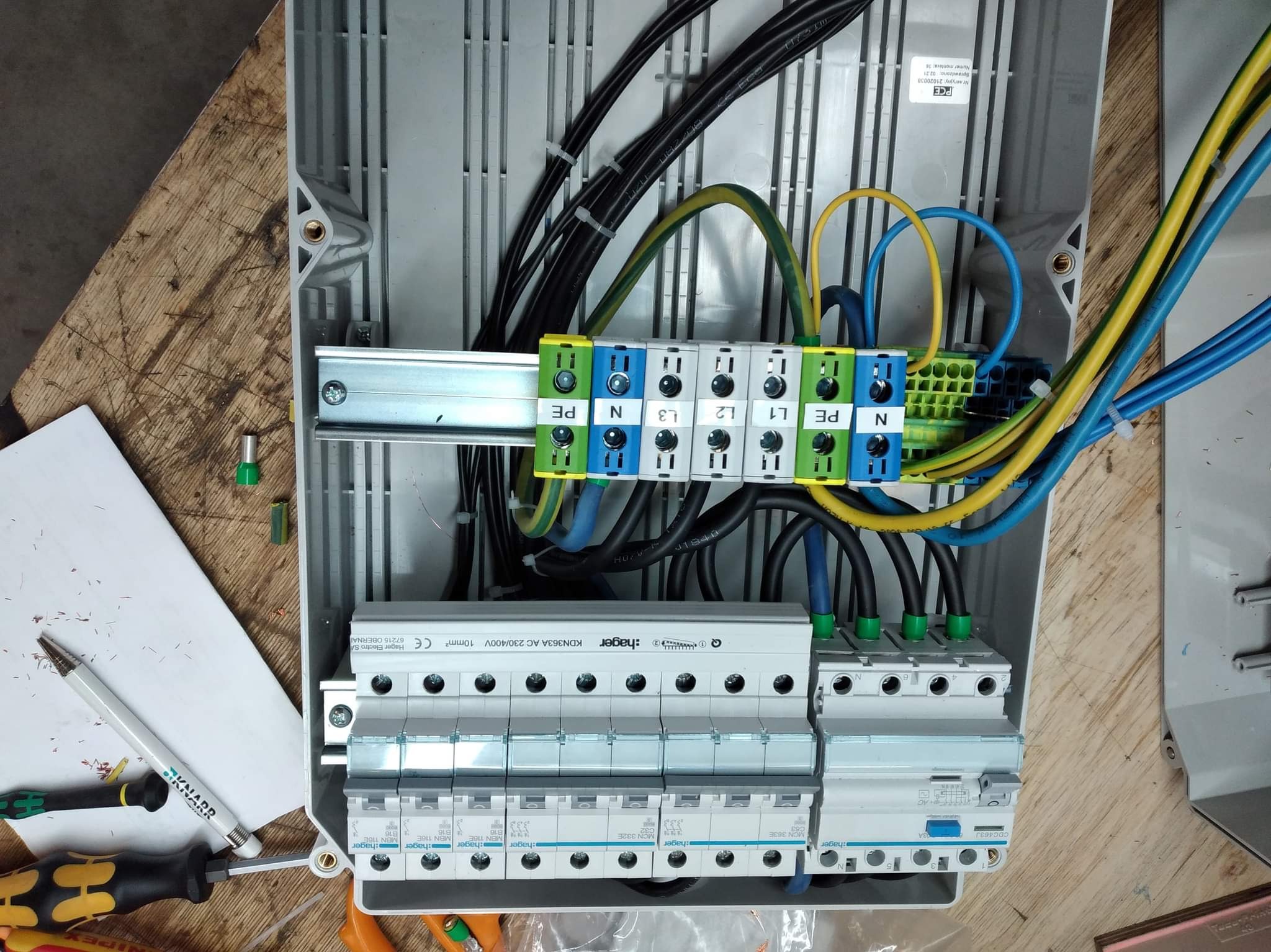 Rozdzielnica elektryczna w trakcie montażu, widoczne zabezpieczenia różnicowoprądowe Hager, listwy zaciskowe PE, N, L1, L2, L3, przewody w izolacji niebieskiej, żółto-zielonej i czarnej, narzędzia...