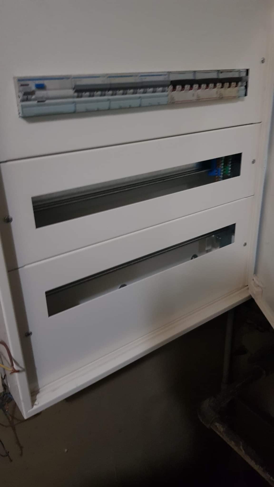 Otwarta, biała skrzynka rozdzielcza z widocznymi bezpiecznikami i przewodami wewnątrz, zamontowana na ścianie w pomieszczeniu.