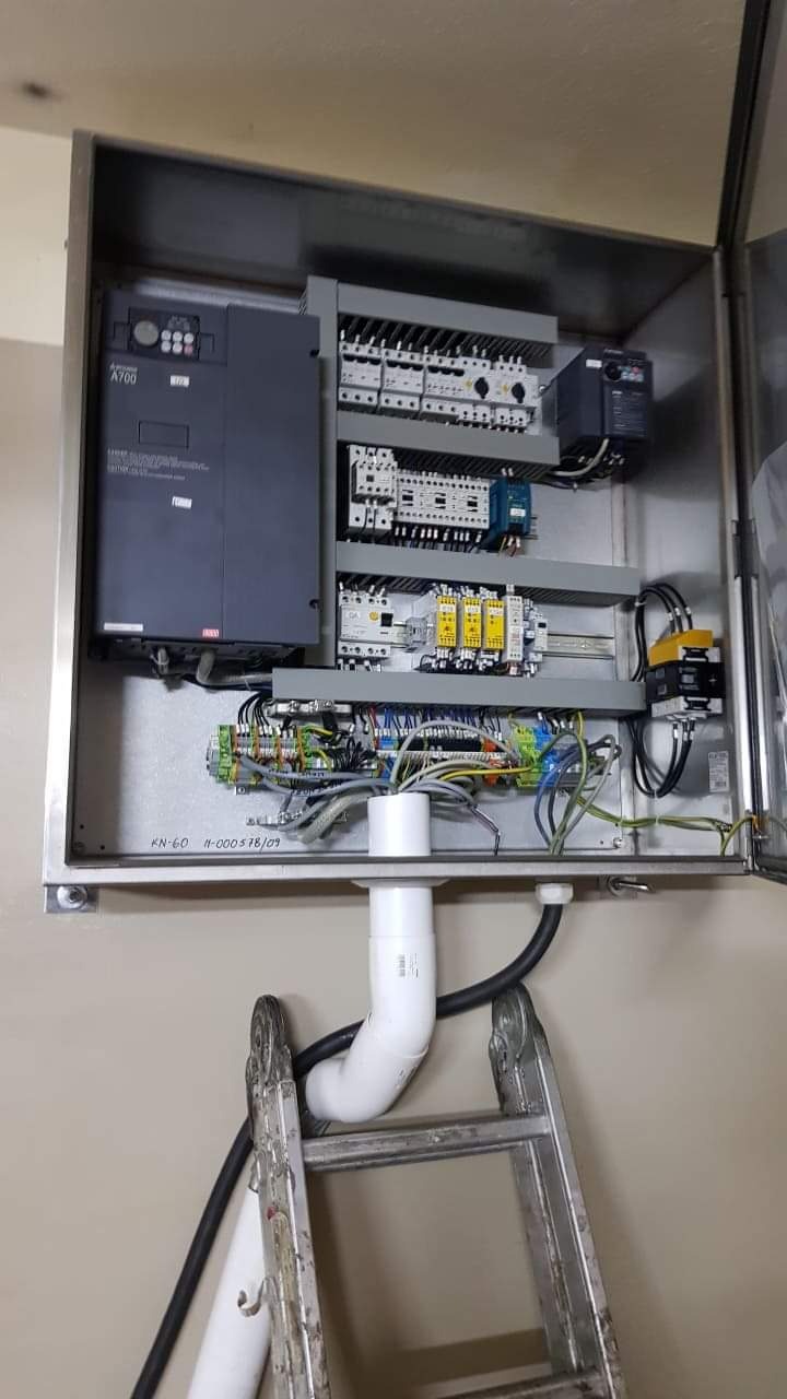 Otwarta metalowa szafa sterownicza z zamontowanym falownikiem Mitsubishi A700, bezpiecznikami, przekaźnikami i okablowaniem, widoczna drabina aluminiowa.