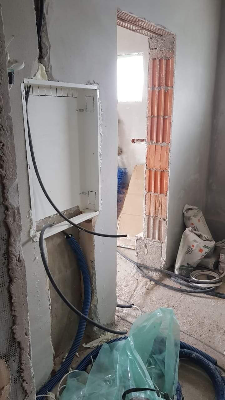Wnętrze w trakcie remontu: pusta biała skrzynka elektryczna w ścianie z wystającymi przewodami i niebieskimi rurkami instalacyjnymi, widoczny fragment muru z cegły dziurawki i worki z zaprawą...
