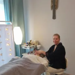 Kobieta wykonuje delikatny masaż twarzy klientce leżącej na łóżku w gabinecie, widoczny wózek z kosmetykami i lampa z dekoracyjnymi światełkami.