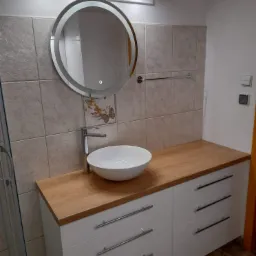 Nowoczesna łazienka z okrągłym lustrem z podświetleniem LED, białą umywalką nablatową na drewnianym blacie i białymi szafkami z trzema rzędami uchwytów.