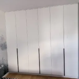 Biała szafa wnękowa z sześcioma drzwiami i długimi, czarnymi uchwytami, na tle ściany z malowanym motywem lasu i ptak&oacute;w, widoczny fragment podłogi z paneli.