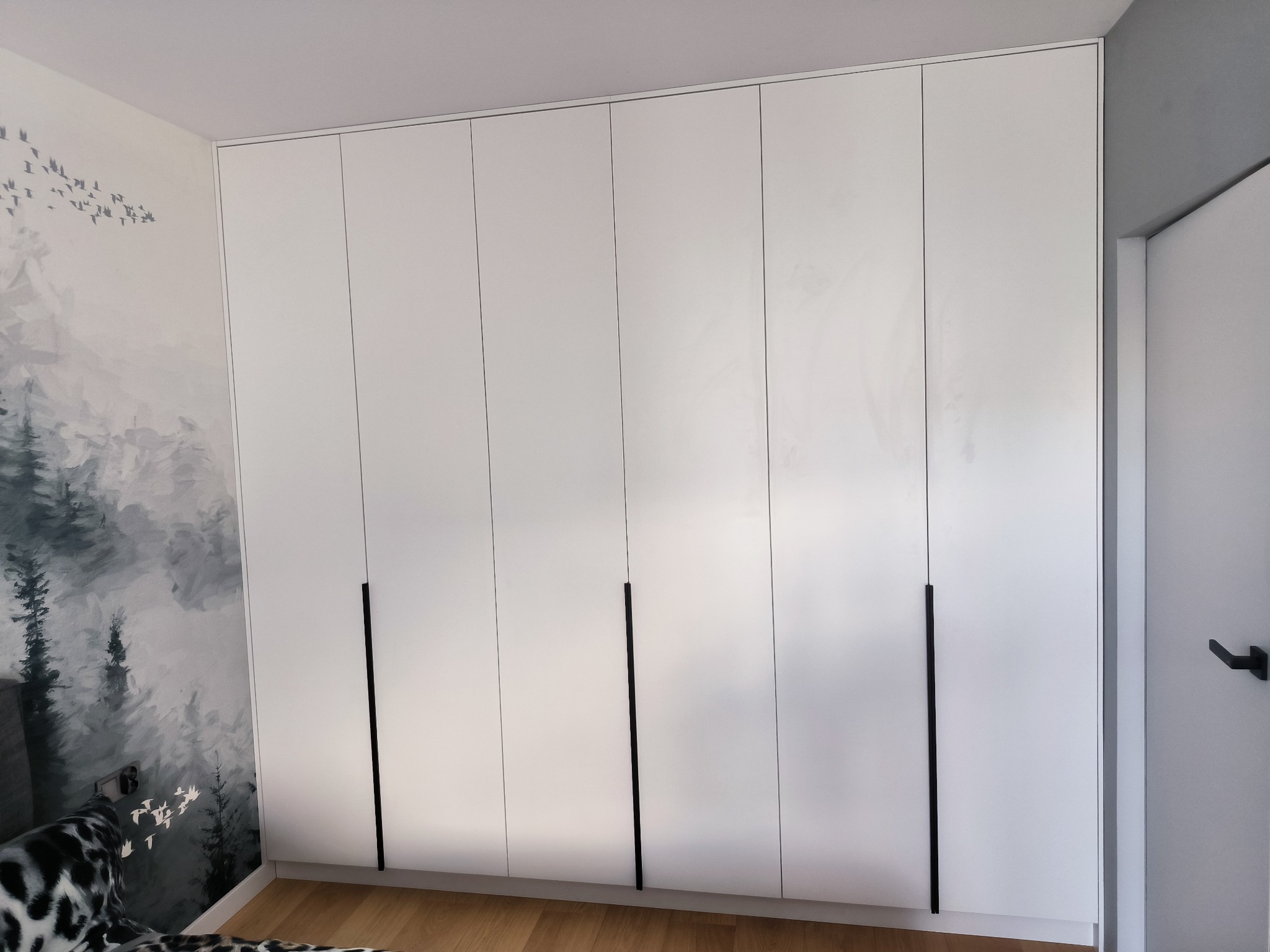 Biała szafa wnękowa z sześcioma drzwiami i długimi, czarnymi uchwytami, na tle ściany z malowanym motywem lasu i ptaków, widoczny fragment podłogi z paneli.
