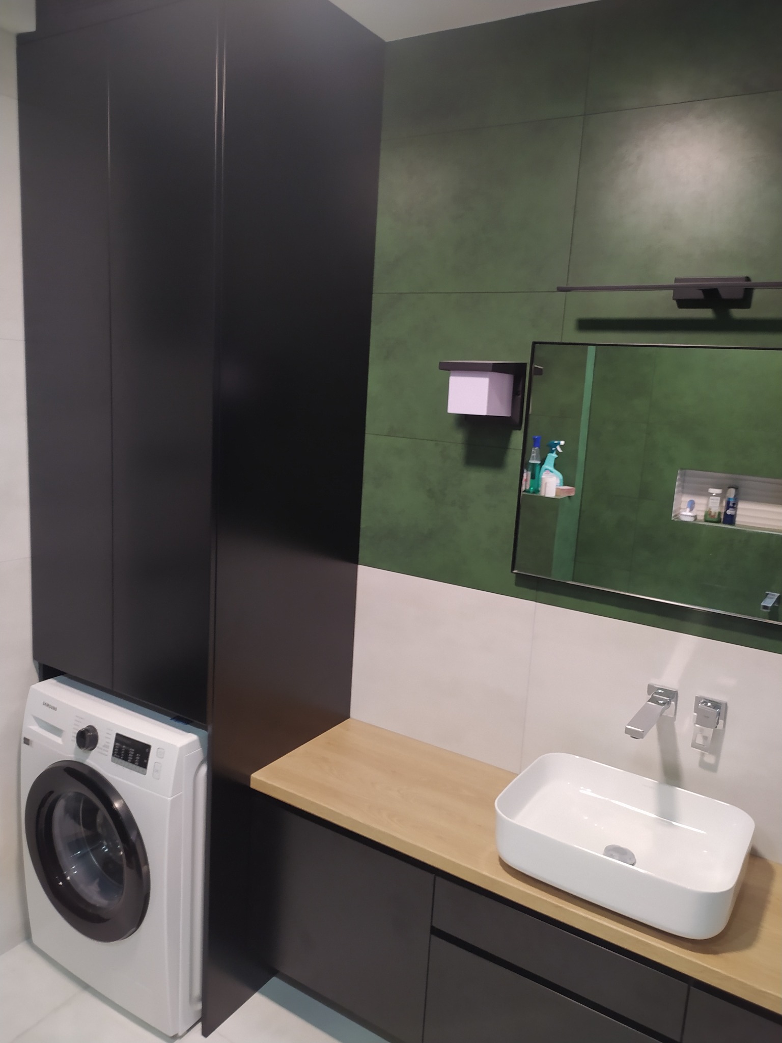 Nowoczesna łazienka z zabudową meblową w kolorze grafitowym, drewnianym blatem, białą umywalką nablatową i pralką Samsung obok szafy, zielona ściana w tle.