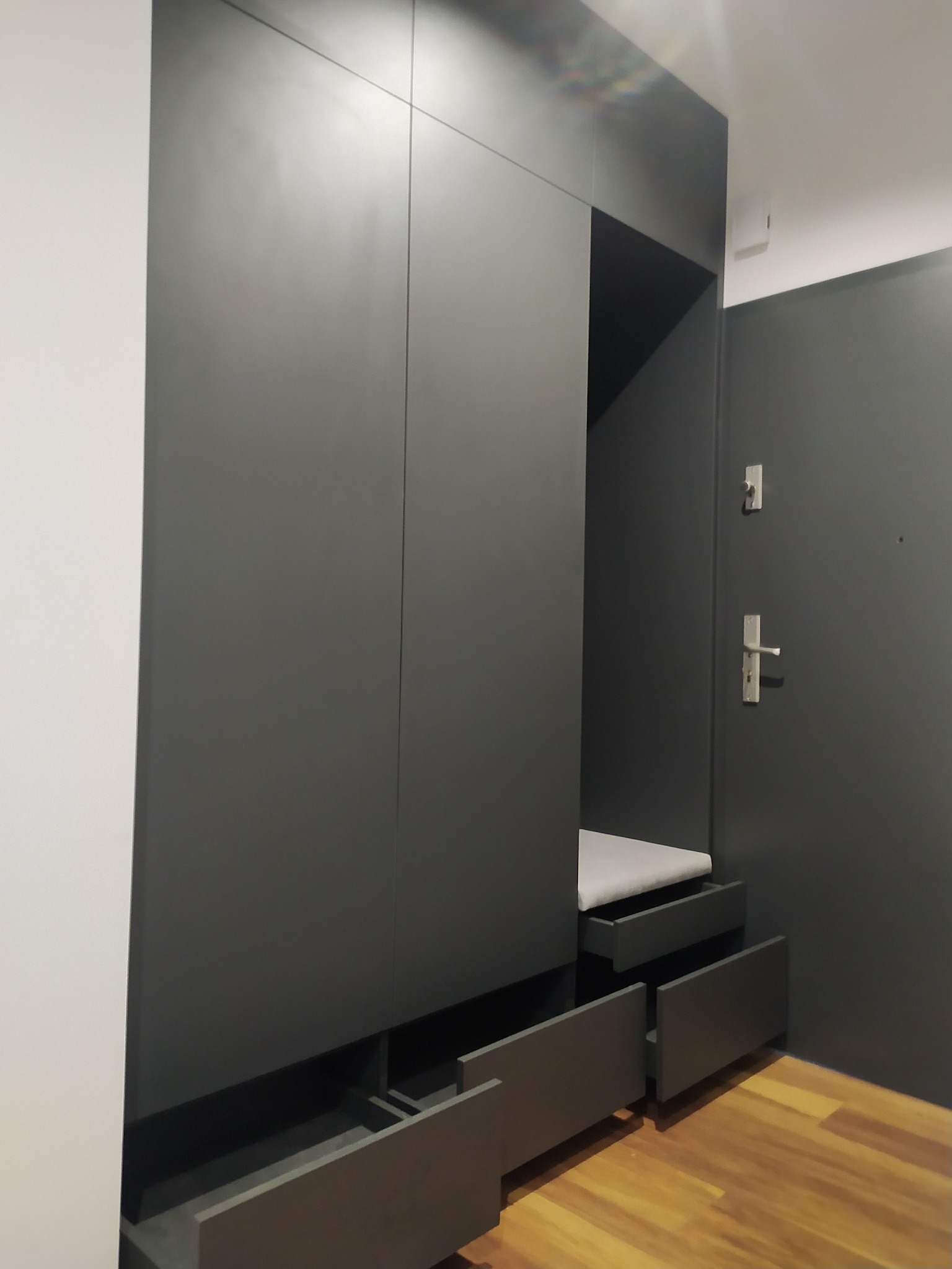 Szara szafa wnękowa z otwartymi szufladami i siedziskiem w przedpokoju, widoczna drewniana podłoga i drzwi wejściowe.