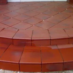 Dwu-stopniowe schody wejściowe wyłożone błyszczącą, czerwoną terakotą, z drobnymi białymi krzyżykami w fugach na górnym poziomie, otoczone ceglaną ścianą i zielenią.