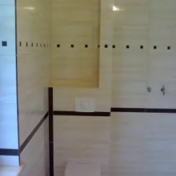 Wykończona łazienka z beżowymi płytkami z ciemnobrązowymi akcentami w postaci cienkich pasków i małych kwadratów, widoczna toaleta i przycisk spłuczki na ścianie.