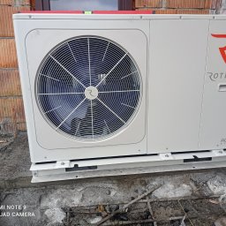 WoleOze.pl - Biała jednostka zewnętrzna z wentylatorem i logo Rotenso zamontowana na betonowej podstawie, widoczna na tle ściany z cegły dziurawki.