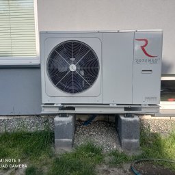WoleOze.pl - Zewnętrzna jednostka pompy ciepła Rotenso Aquami Series zamontowana na betonowych bloczkach z widoczną instalacją i fragmentem trawnika.