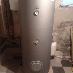 WoleOze.pl - Szary, pionowy zbiornik buforowy Galmet z manometrem, zainstalowany w piwnicy z widocznymi rurami i zbiornikiem wyrównawczym, na tle ściany z plamami wilgoci i szachownicowej podłogi.