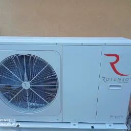 Biała zewnętrzna jednostka z wentylatorem i czerwonym logo 'Rotenso' serii Aquami, zamontowana na zewnątrz budynku z beżową elewacją, widoczny fragment metalowego ogrodzenia.