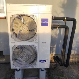 WoleOze.pl - Zewnętrzna jednostka klimatyzacyjna Haier z dwoma wentylatorami, zamontowana na betonowych bloczkach przy jasnej ścianie, widoczne czarne rury izolacyjne z mosiężnymi złączkami.