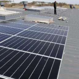 Montaż paneli słonecznych na dachu przemysłowym, widoczne ułożone panele, pracownicy instalujący system, elementy montażowe i wentylacja dachowa.