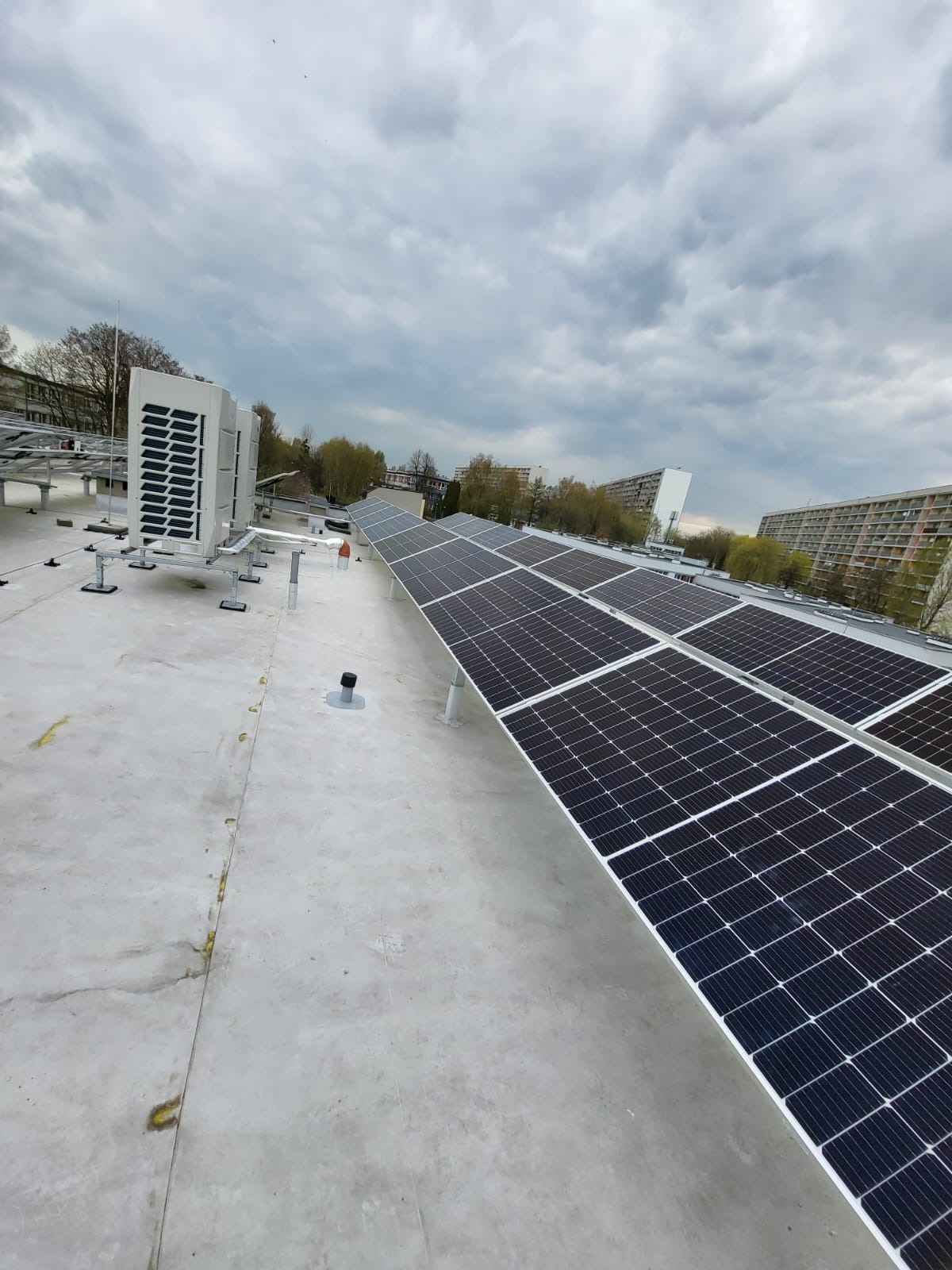Instalacja paneli słonecznych na płaskim dachu budynku, widoczne również jednostki zewnętrzne klimatyzacji i zabudowa miejska w tle, pochmurne niebo.
