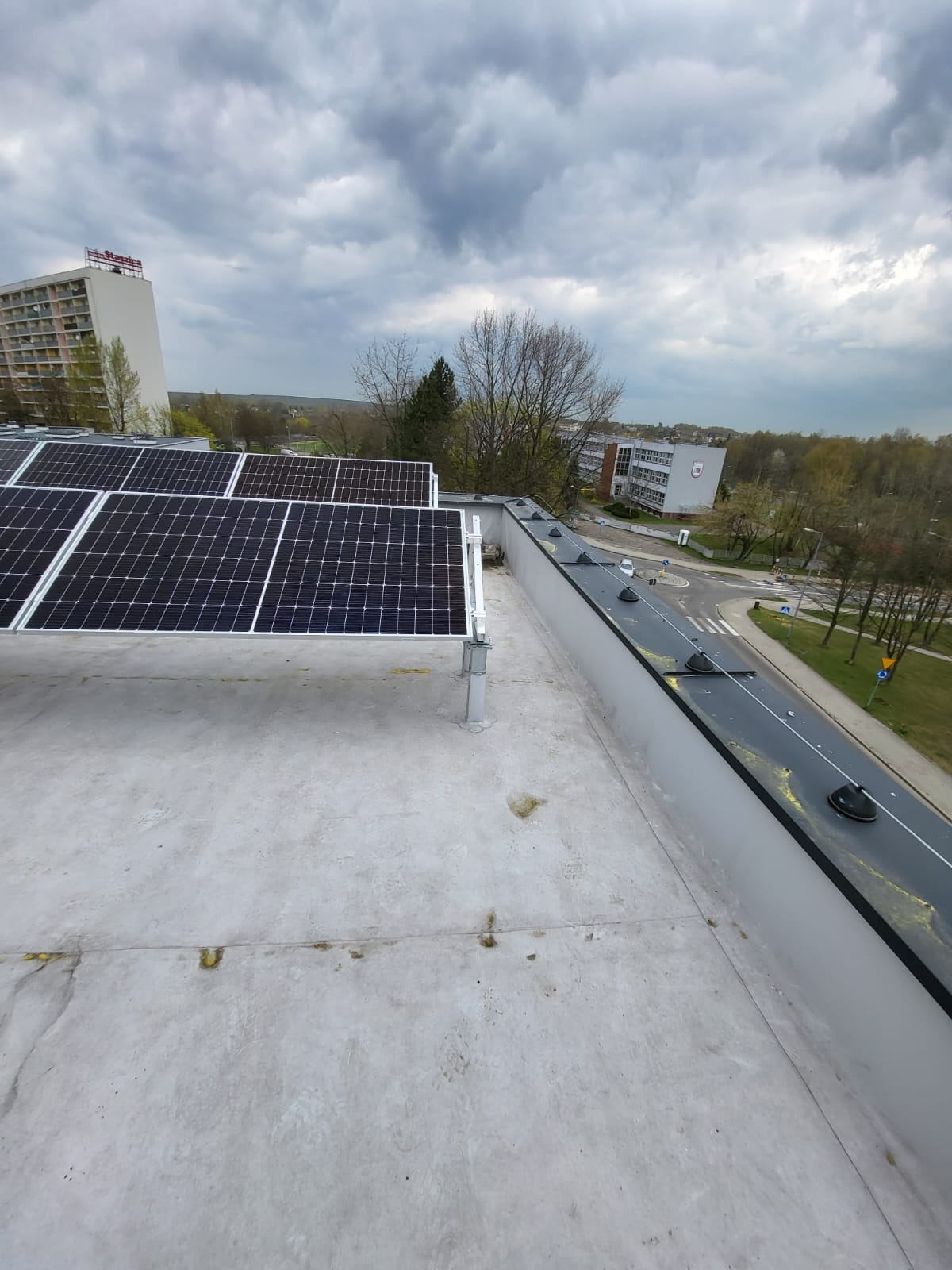 Panele fotowoltaiczne zamontowane na płaskim dachu budynku, widok z góry na instalację i otoczenie miejskie w pochmurny dzień.