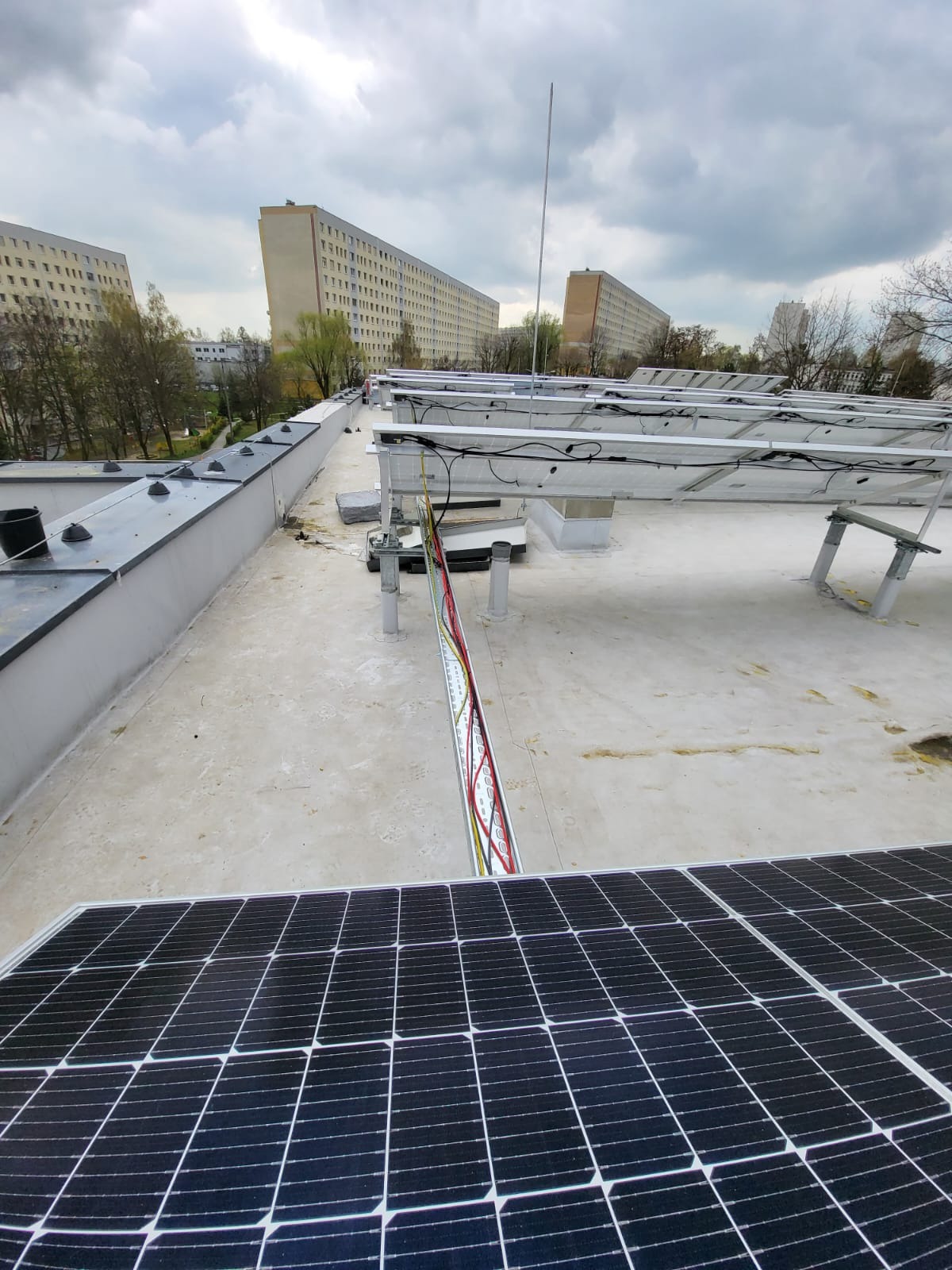 Montaż paneli słonecznych na płaskim dachu bloku mieszkalnego, widoczne kable w korytku, elementy konstrukcji wsporczej i fragmenty innych paneli w tle.