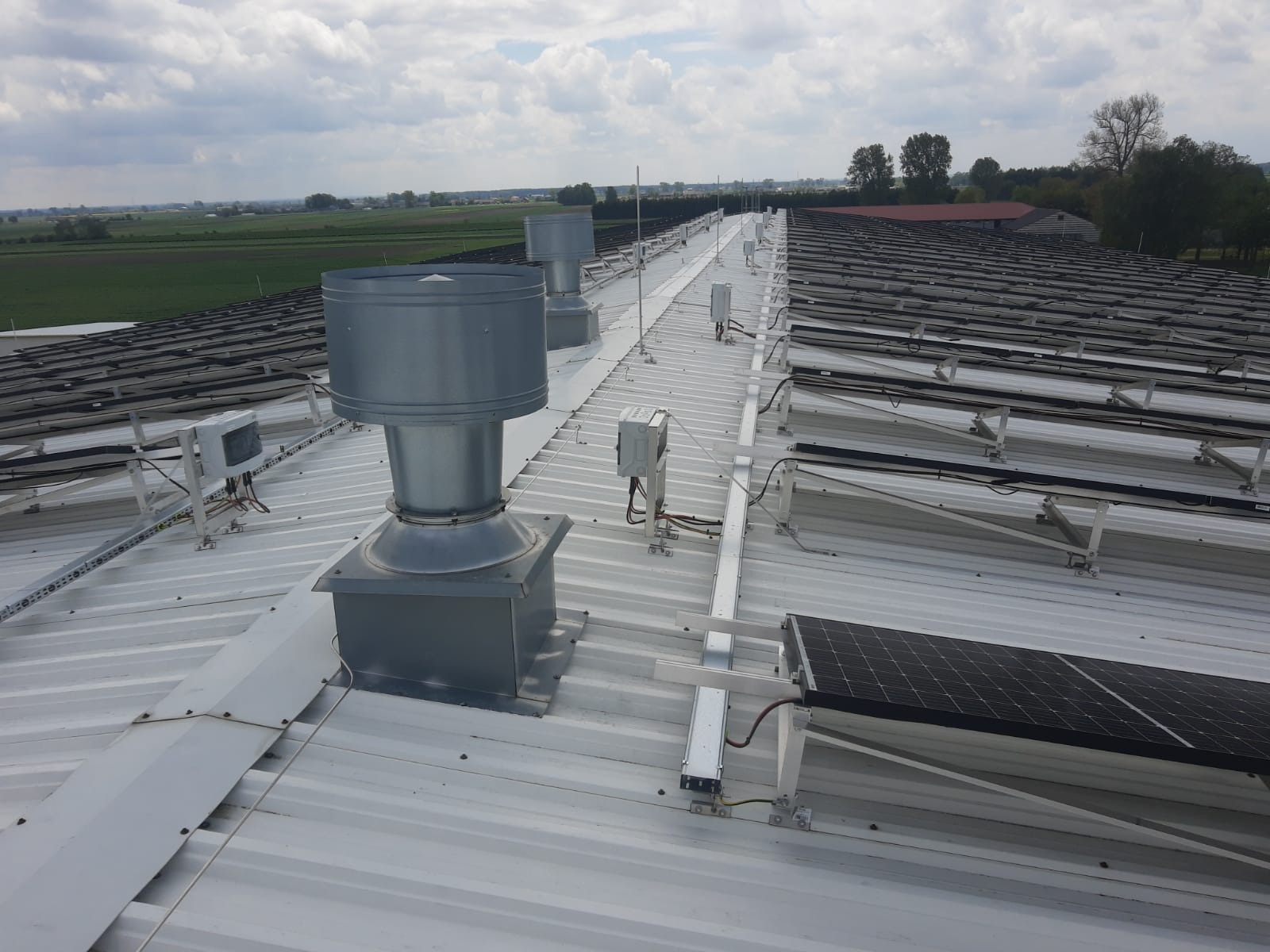 Instalacja fotowoltaiczna na dachu hali przemysłowej z widocznymi panelami słonecznymi, systemem montażowym, przewodami oraz elementami wentylacji. W tle rozległe pola i lasy pod pochmurnym niebem.