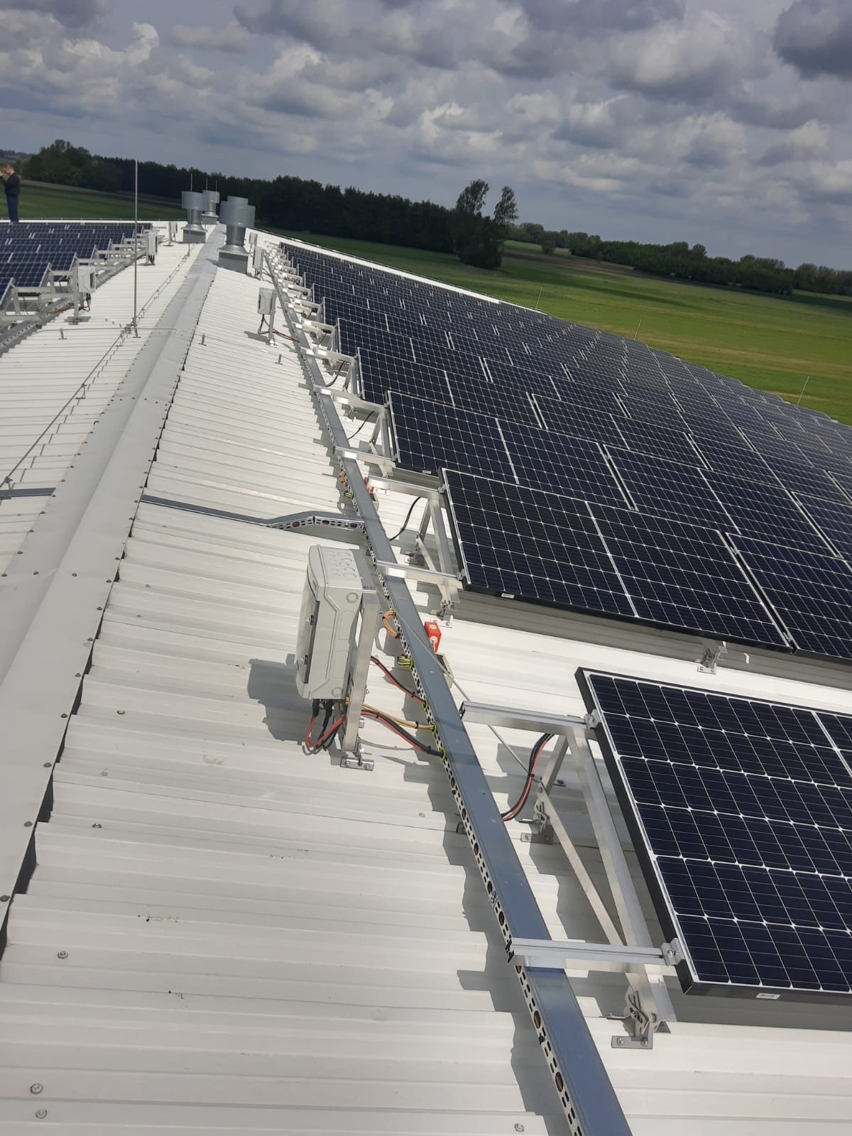 Rząd paneli słonecznych zamontowanych na metalowym dachu budynku, z widocznym inwerterem i systemem mocowań, na tle zielonych pól i pochmurnego nieba.