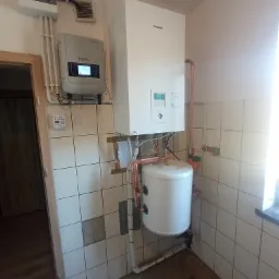 Biały piec gazowy Panasonic z zasobnikiem ciepłej wody, połączony miedzianymi rurami, zamontowany w pomieszczeniu z wyłożonymi kafelkami ścianami, obok widoczny inwerter solarny MASS SOLAR.