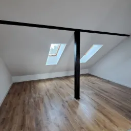 Jasne poddasze z drewnianą podłogą i dwoma oknami dachowymi. Czarne belki stropowe i filar dodają kontrastu. Świeżo wykończone wnętrze, gotowe do aranżacji.