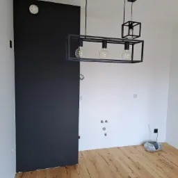 Wnętrze po remoncie: czarna ściana, drewniana podłoga, nowoczesne lampy wiszące z żarówkami Edisona, widoczne gniazdka elektryczne i instalacja wodna, radio na podłodze.