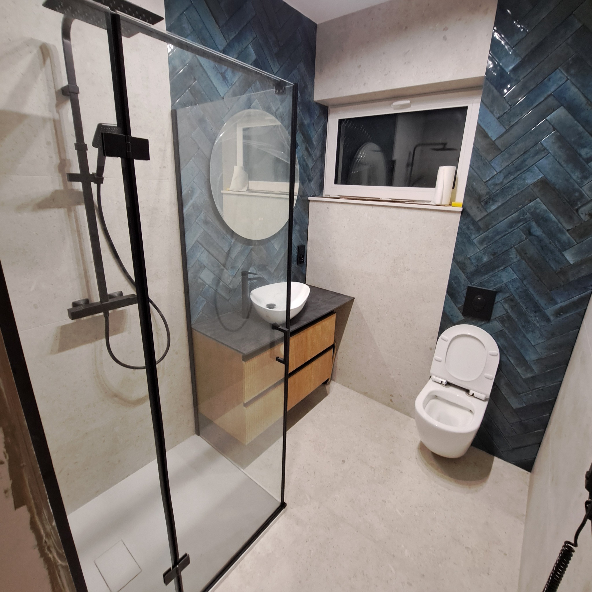 Nowoczesna łazienka z kabiną prysznicową typu walk-in z czarną ramą, umywalką nablatową na drewnianej szafce z szufladami i ścianą wyłożoną niebieską glazurą ułożoną w jodełkę.
