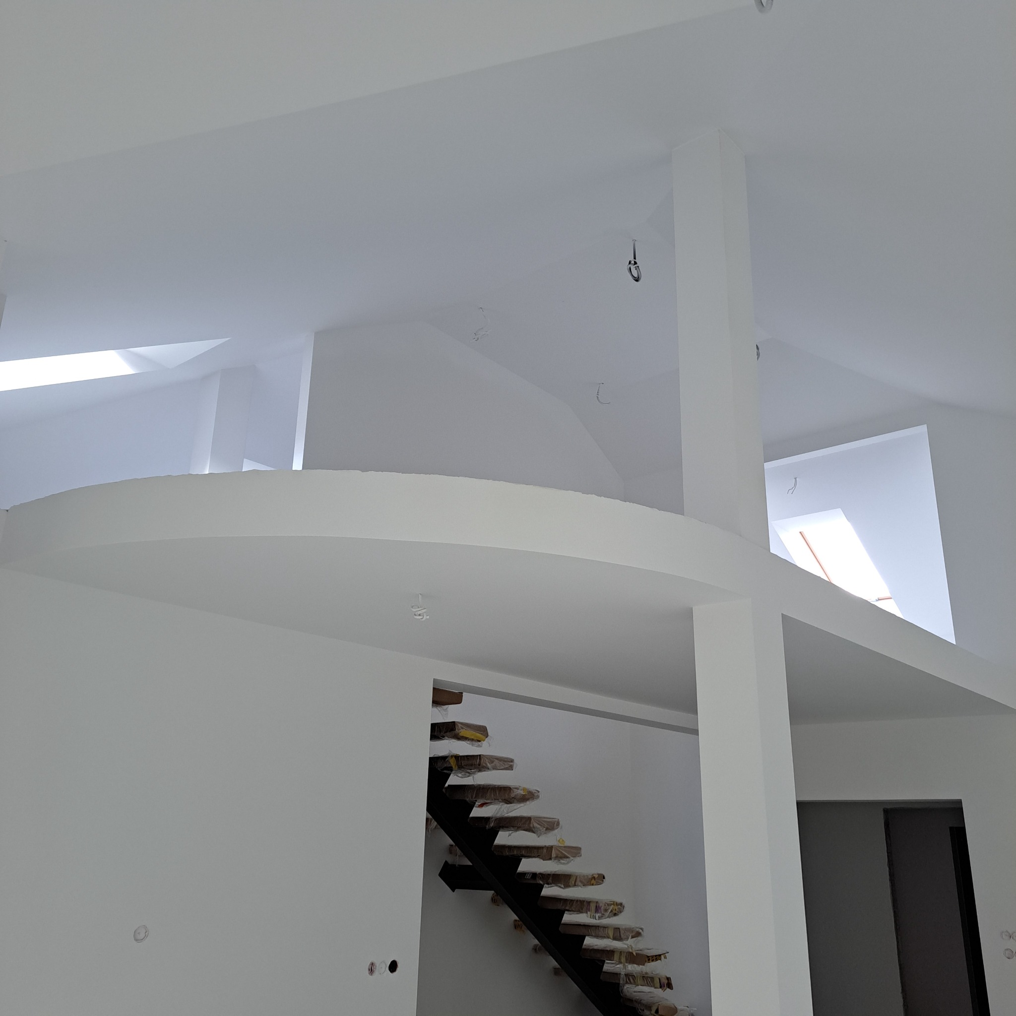 Białe, przestronne wnętrze domu po malowaniu, z widocznymi schodami zabezpieczonymi folią, antresolą i oknami dachowymi. Minimalistyczny design.