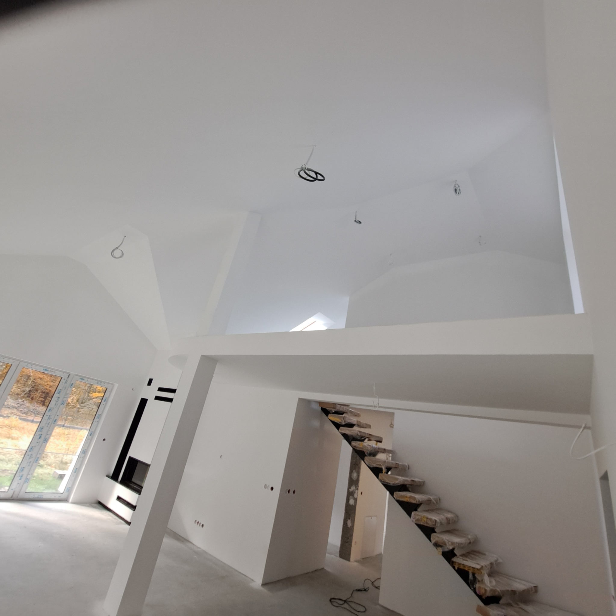 Wnętrze domu po remoncie: białe ściany, schody z zabezpieczeniami, okno z widokiem na drzewa. Widoczne punkty oświetleniowe na suficie.